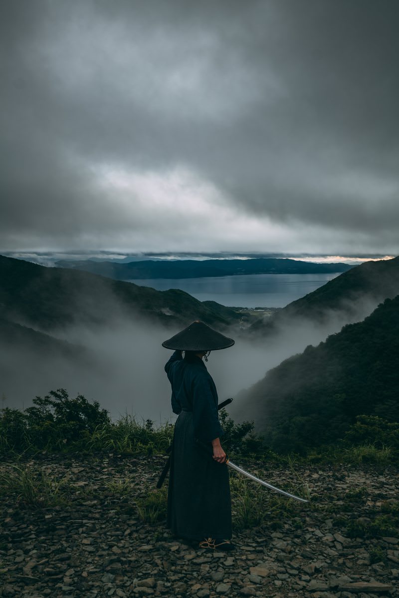 灰色の雲が立ち込める峠道で刀を抜いて構える笠をかぶった侍［モデル：まーこ］