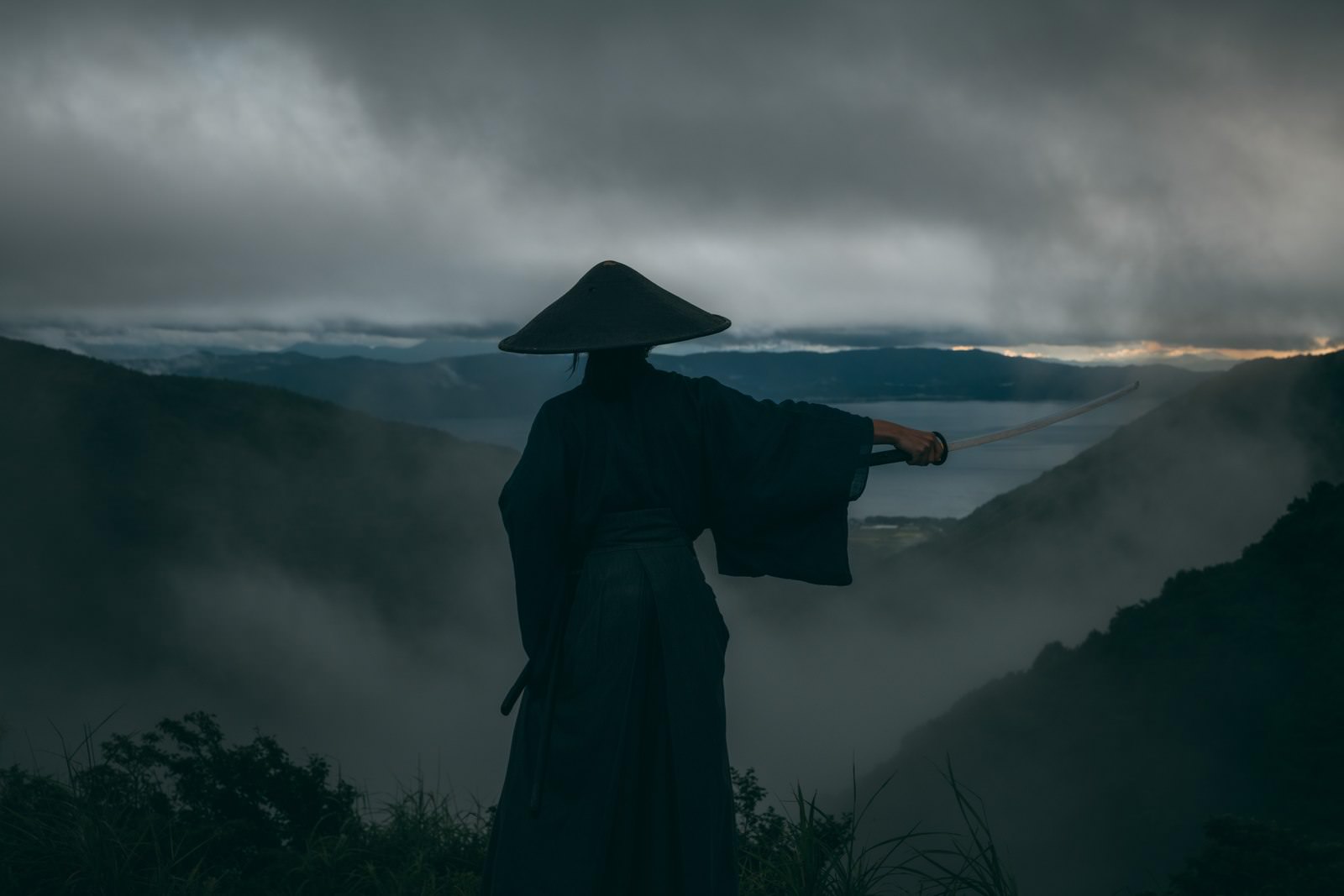 重い雲が立ち込める中、網代笠を被った侍が日本刀を構える姿［モデル：まーこ］