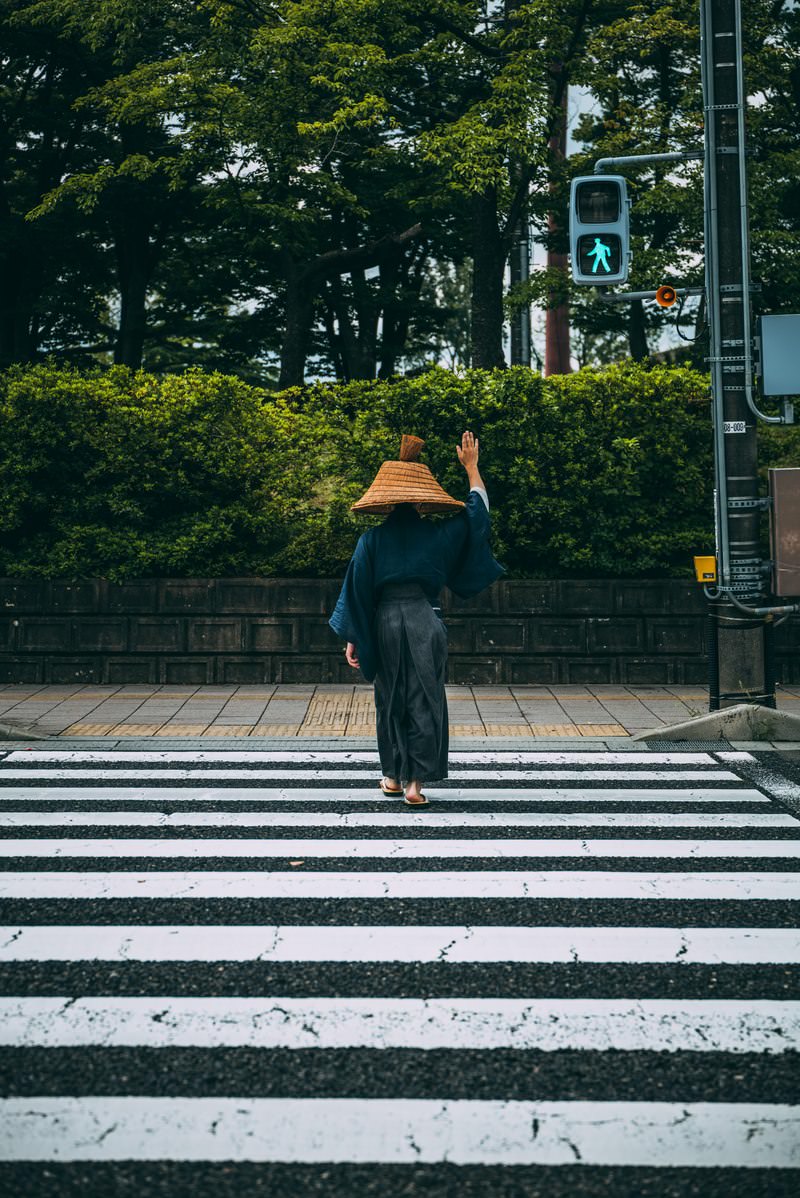 網代笠をかぶった侍が手を上げて横断歩道を渡っている様子［モデル：まーこ］