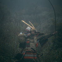 藪の中で大弓を引く大鎧姿の武士が弓矢を放つ瞬間の写真