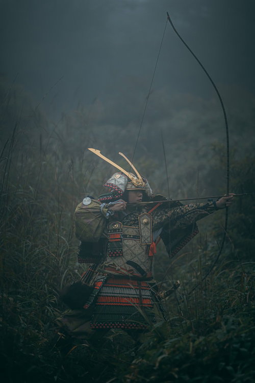 藪の中で大弓を引く大鎧姿の武士が弓矢を放つ瞬間