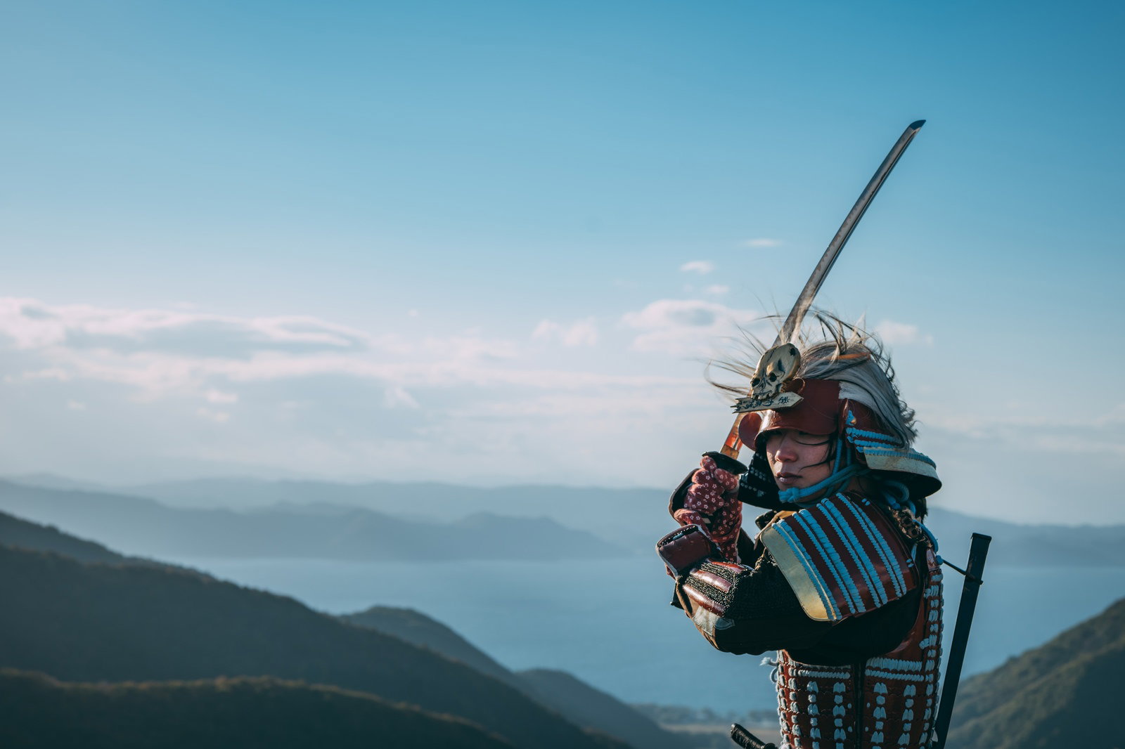 山を背景に上段の構えで日本刀を構える甲冑姿の武士［モデル：まーこ］