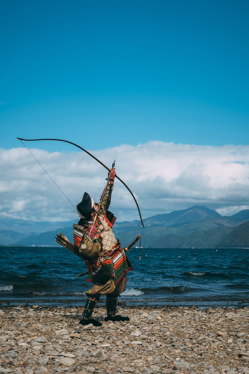 湖畔の山々を背景に空に向かって大弓を引く武士