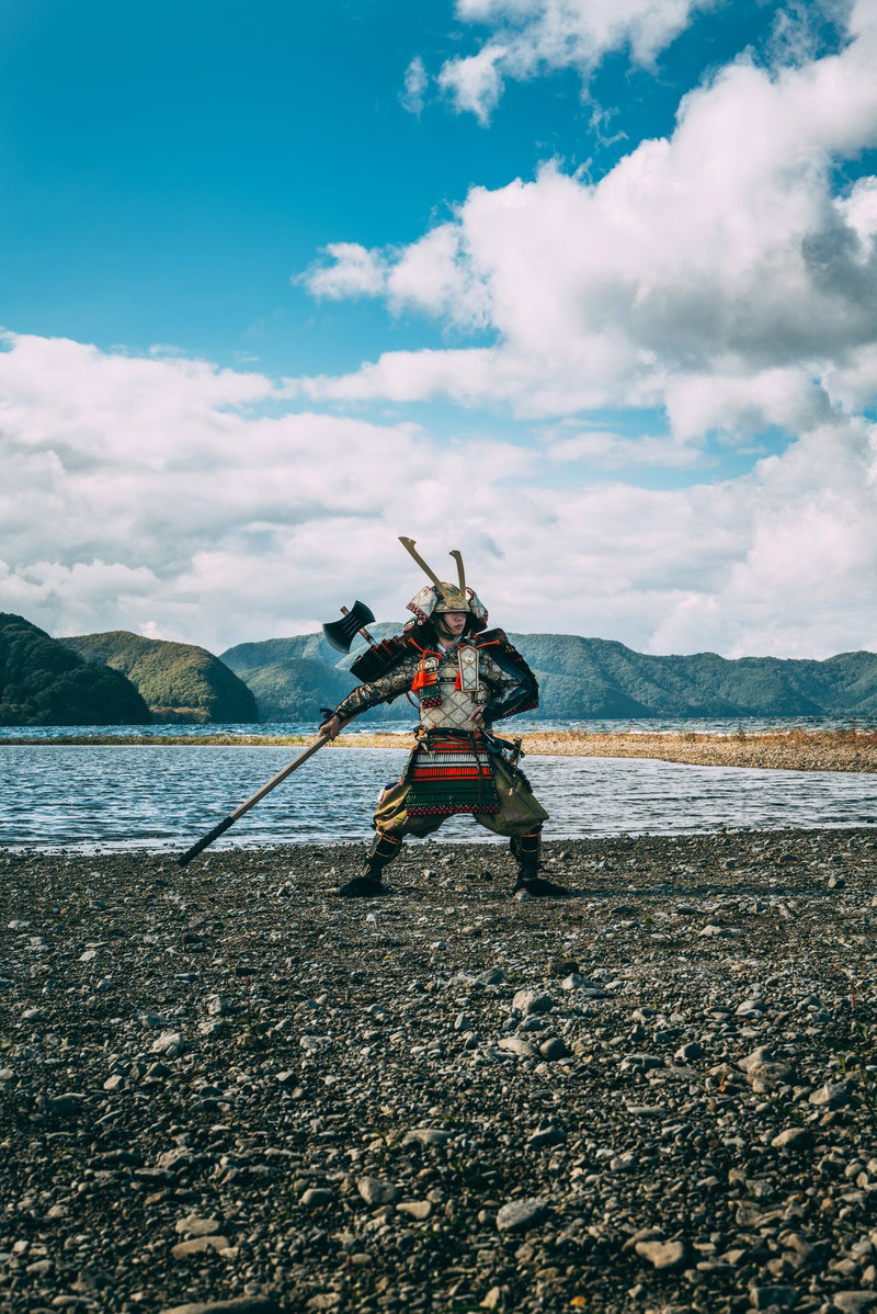 海辺の砂浜で棍棒を振りかぶる黒い大鎧の武将［モデル：まーこ］
