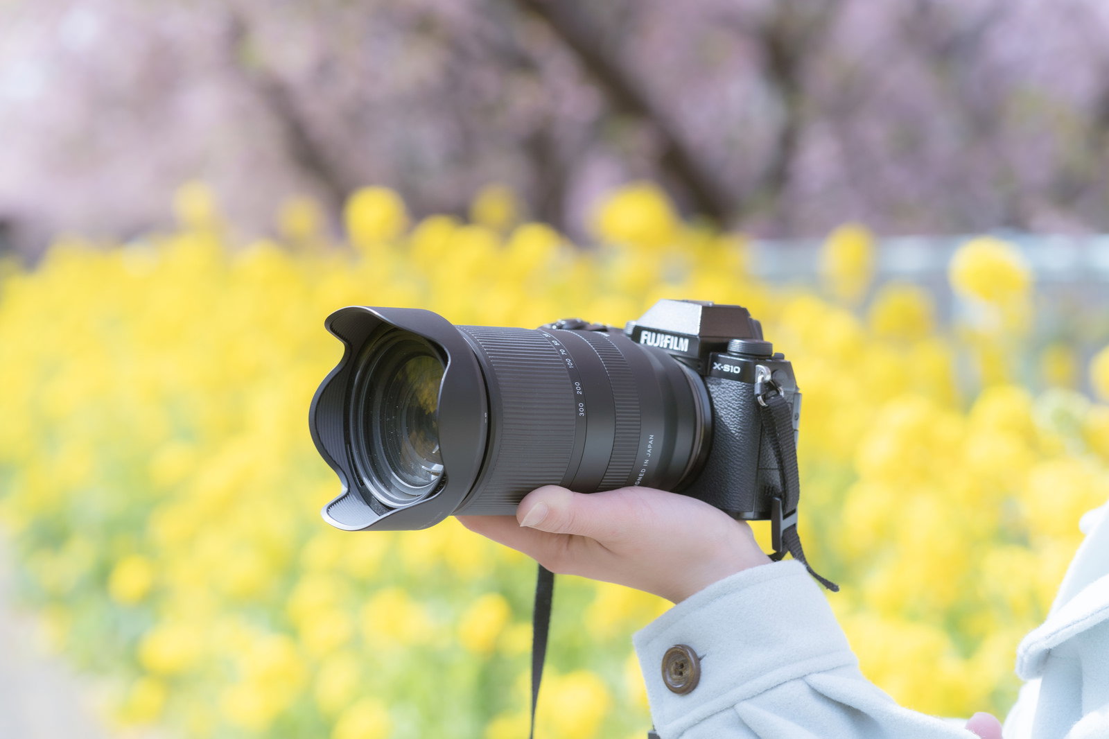 手の上に乗せた大口径レンズ付き一眼カメラ、背景に菜の花