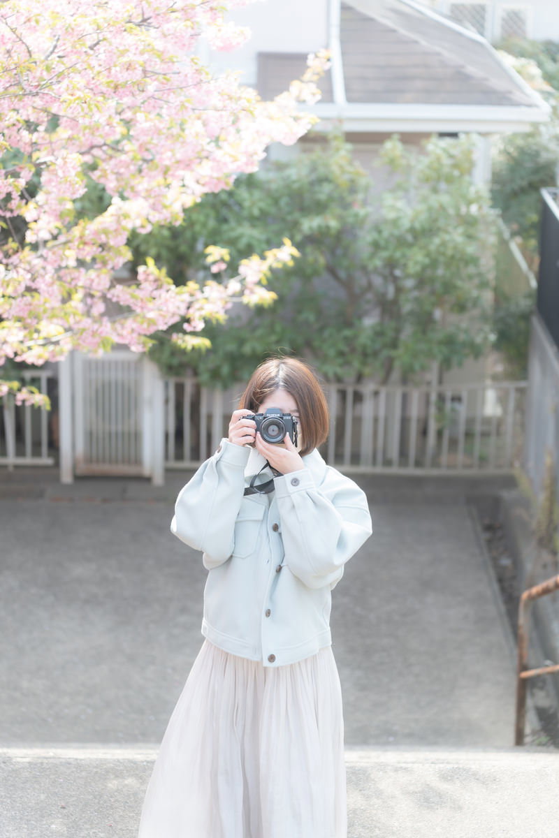 石段でカメラを構える白いコートの女性、桜が咲く春の風景