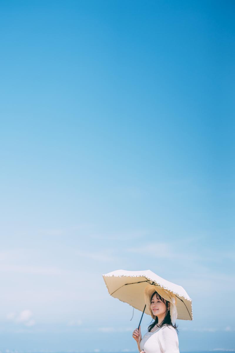 青空を背景に、ベージュの日傘を持ち上を向く女性のポートレート写真［モデル：茜さや］