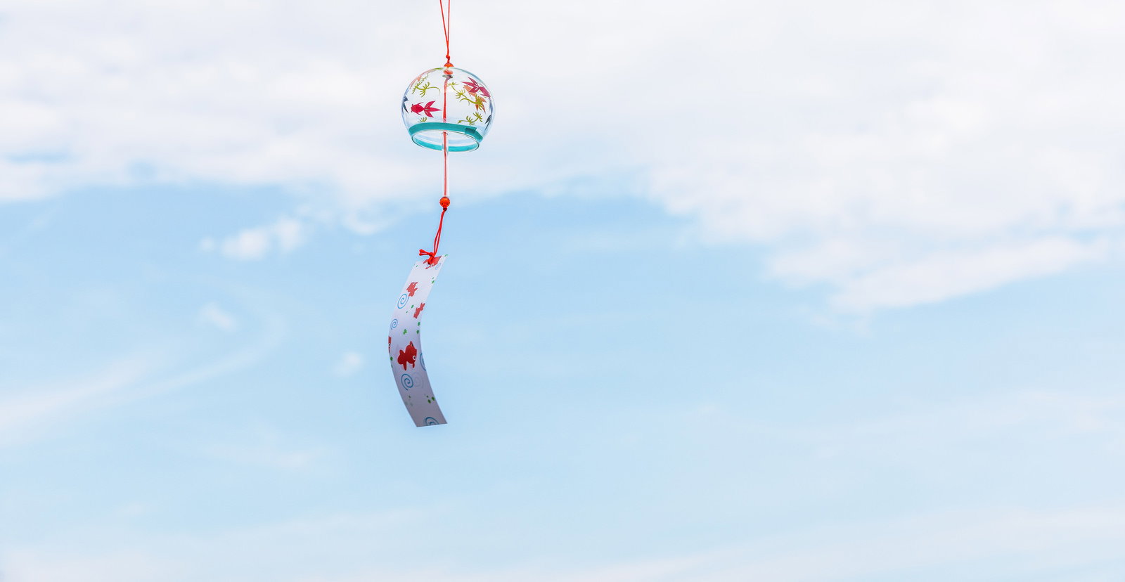青い空を背景に、赤い紐で吊られた花柄の透明な風鈴が揺らいでいる