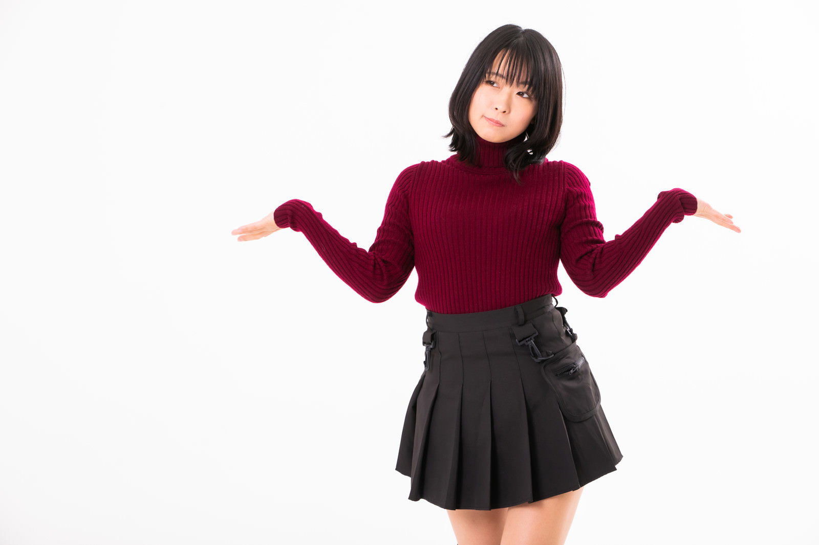 エンジ色のニットと黒いプリーツスカートを着た女性が両腕を上げてお手上げポーズをしている［モデル：にゃるる］