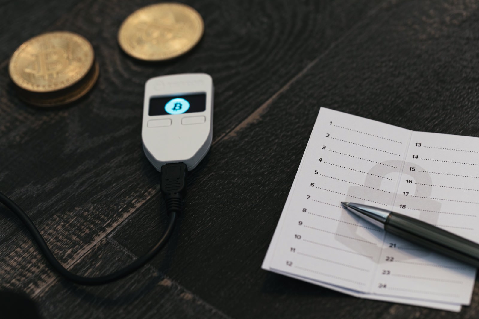 黒いテーブルの上に配置されたTREZORハードウェアウォレット、ペン、メモ用紙、ビットコインのコイン