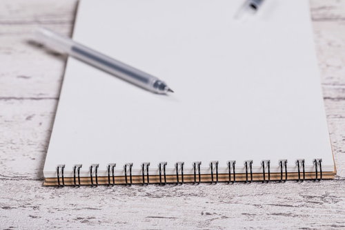 木目調のデスクに置かれた何も書かれていないメモ用紙とペン