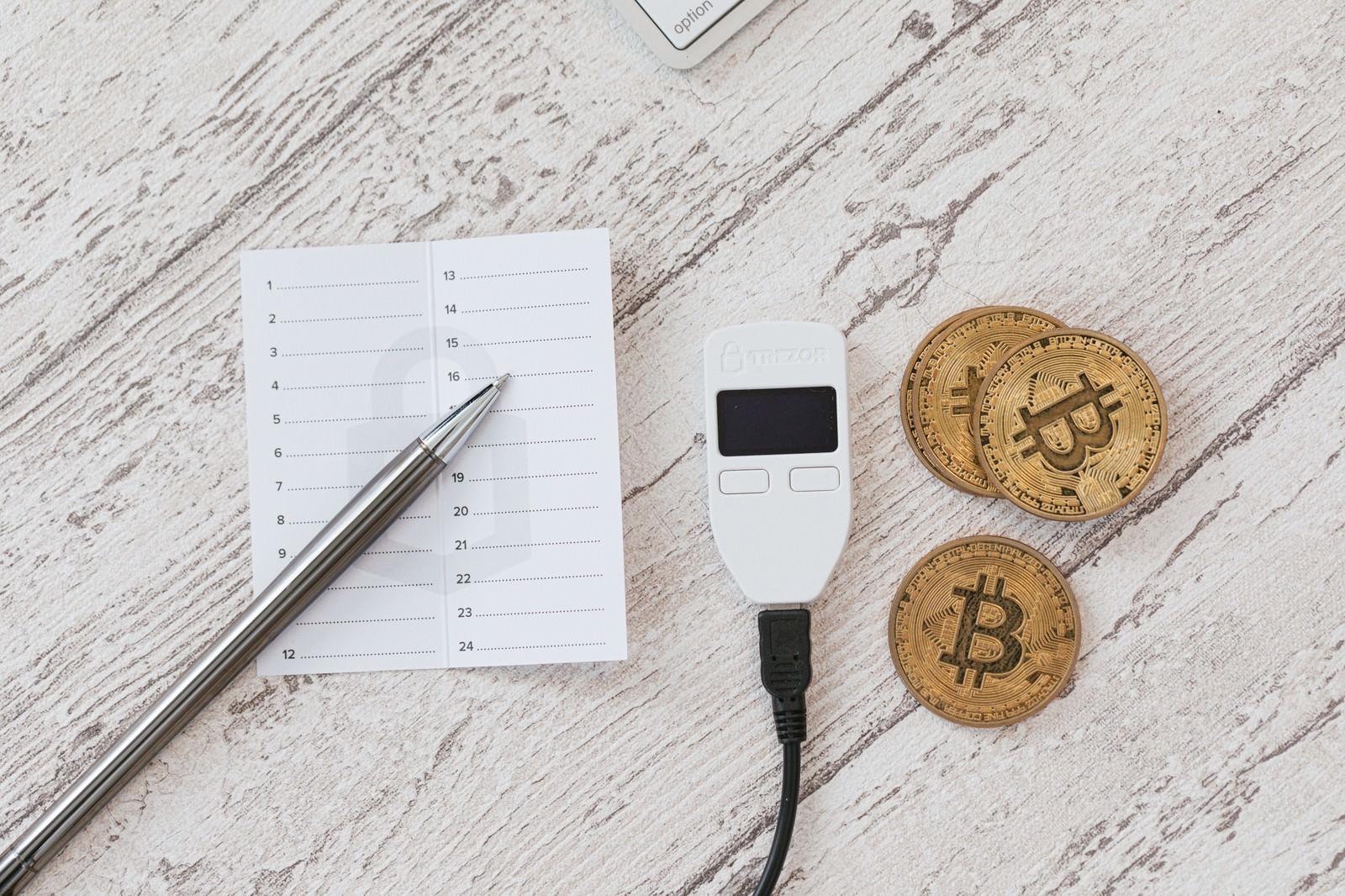 白いメモ用紙とペン、黒いTREZORウォレット、金色のビットコインコインが木目調の背景に並べられている