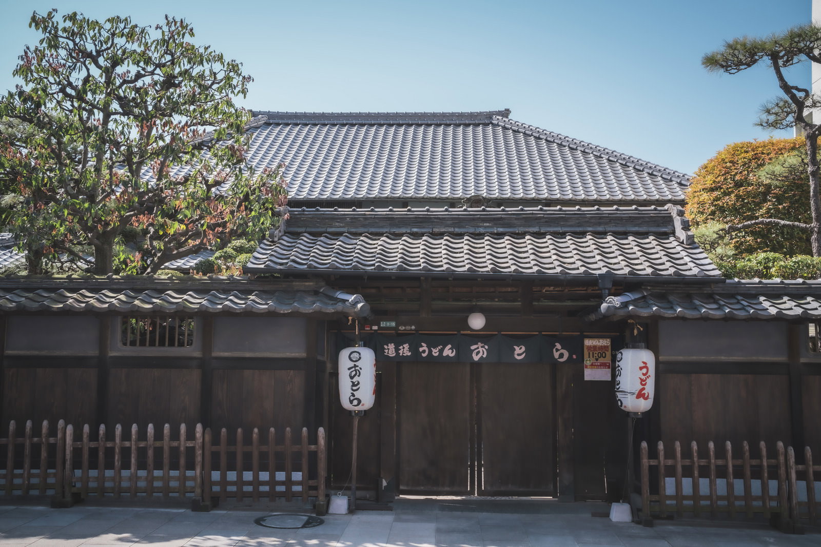 青い瓦屋根の黒い薬医門と白い木製の塀、提灯が見える伊予食堂おとらの門構え