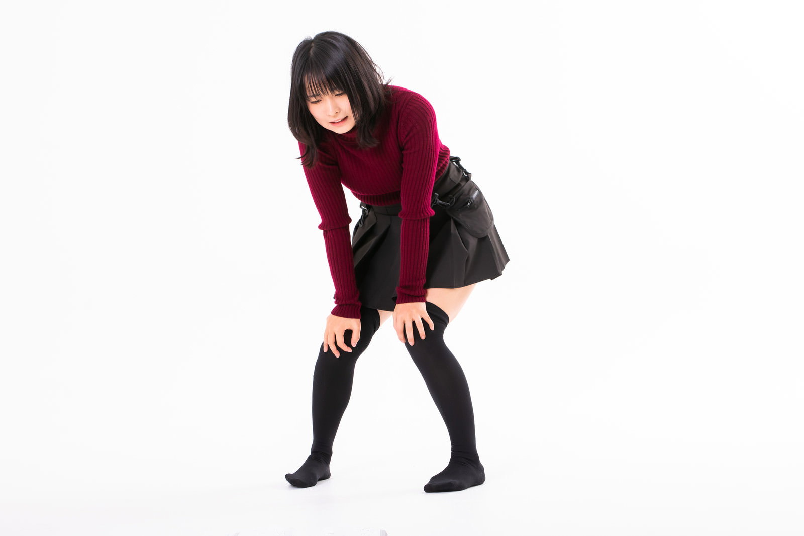 赤いニットを着た女性が膝に手をあてて疲れた表情で立っている［モデル：にゃるる］