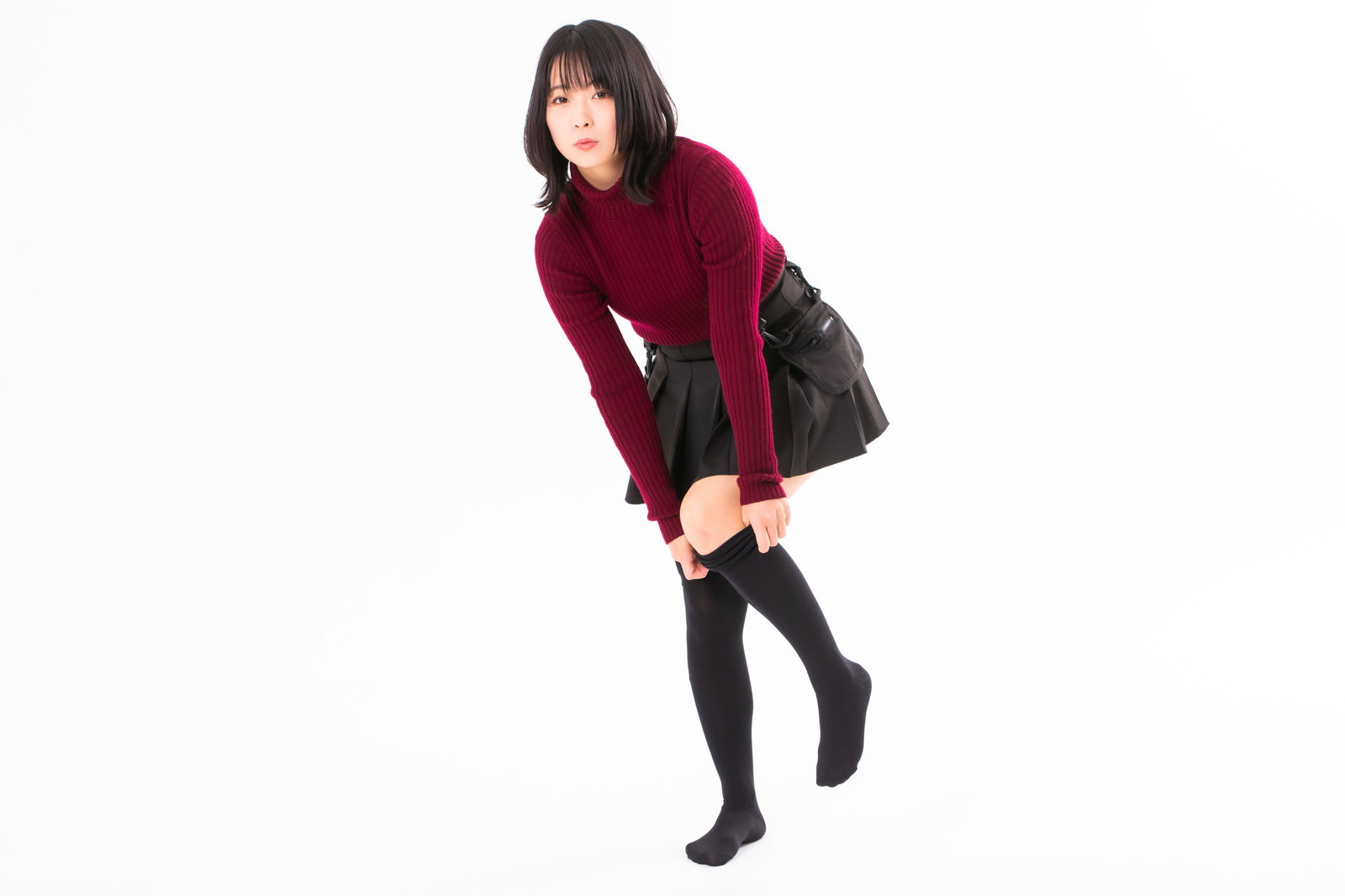 白背景で赤いトップスと黒いスカートを着て、ハイソックスを引っ張り上げる女性［モデル：にゃるる］
