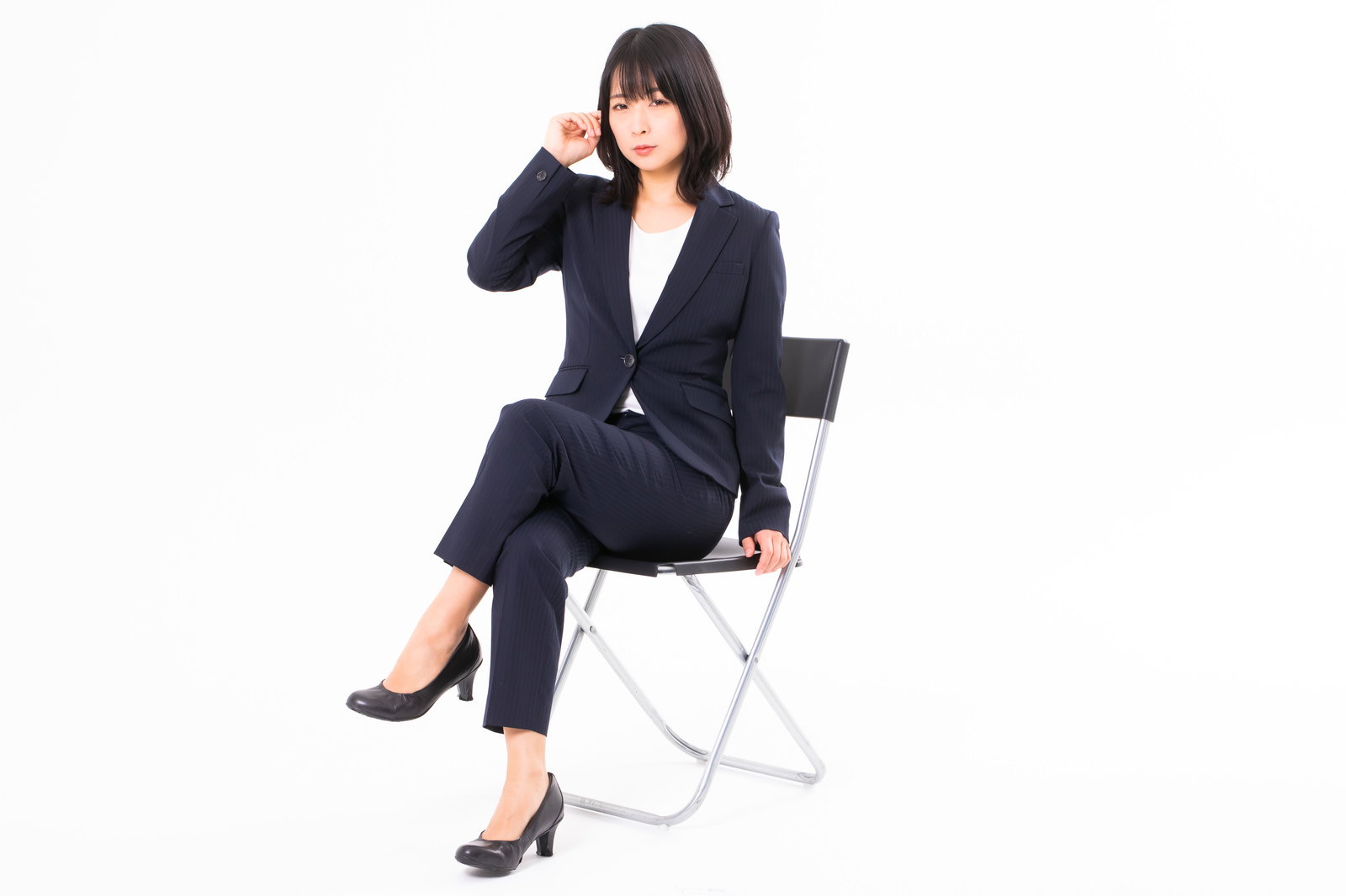 黒いスーツを着た女性が白い椅子に腰かけて足組みしている［モデル：にゃるる］