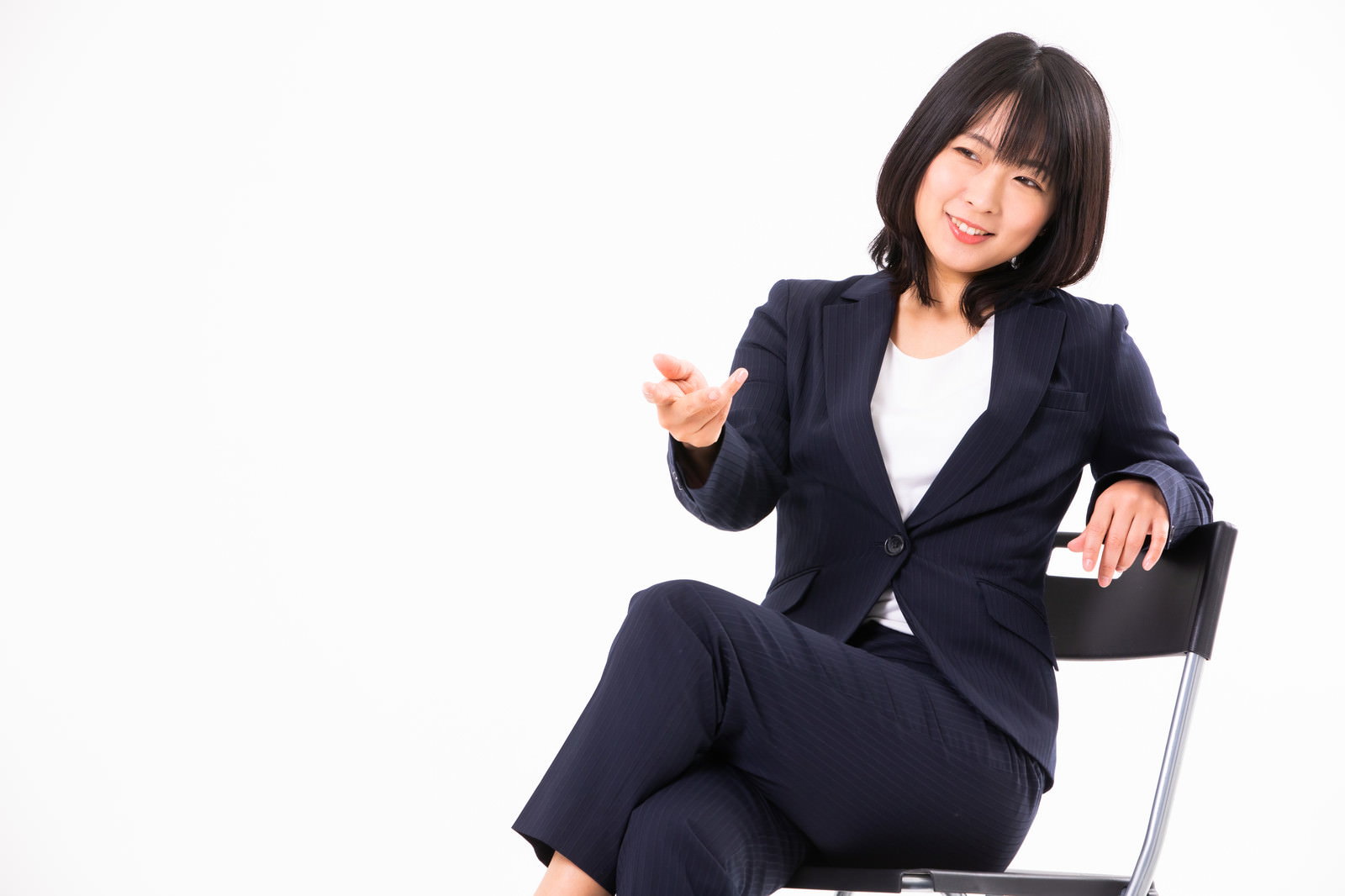 椅子に座って片手を上げながらざっくばらんに話している紺色スーツの女性社員［モデル：にゃるる］