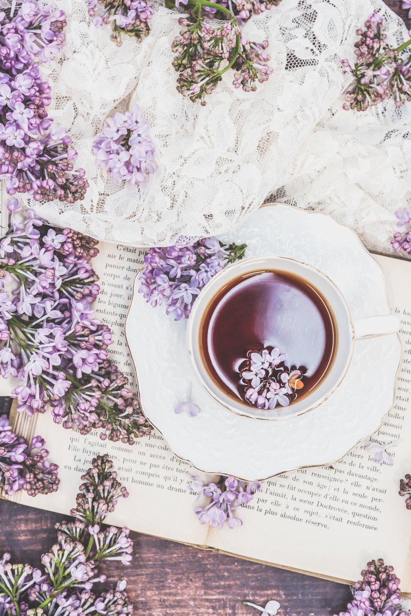 白いティーカップに入ったハーブティーにライラックの花が浮かび、レースと洋書が配置された写真