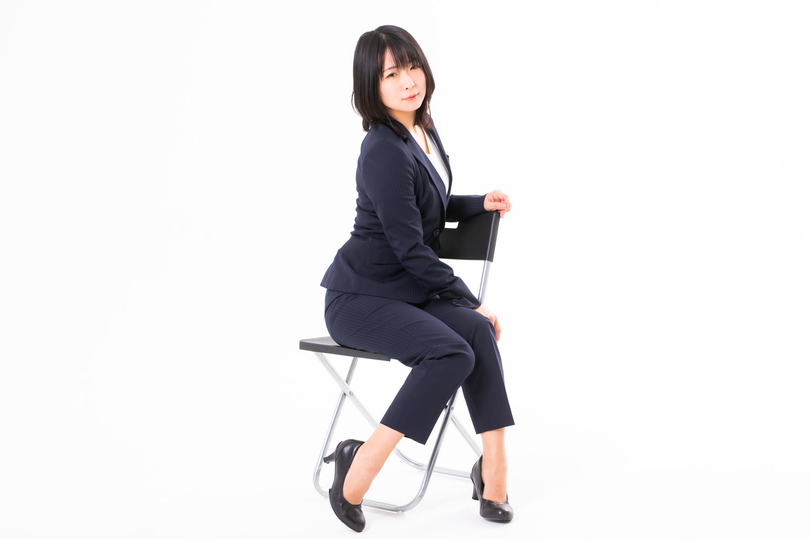 紺色のスーツを着た女性社員が白い椅子に腰かけて振り返る様子［モデル：にゃるる］