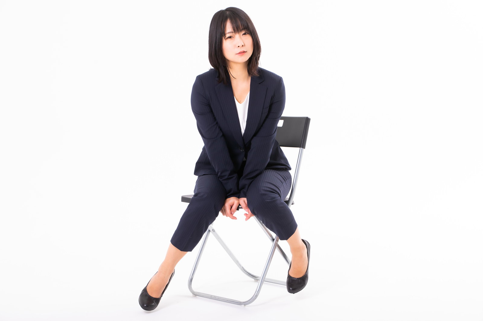 黒いスーツを着た女性が白い椅子に腰掛け、両手を膝の間に挟んでいる［モデル：にゃるる］