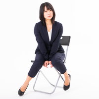 黒いスーツを着た女性社員が白い椅子に腰掛け両手を膝に挟み込むポーズの写真