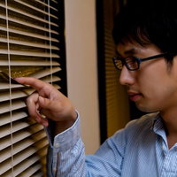 暗い室内でブラインドの隙間から外を覗く男性の写真