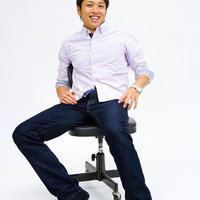 白い背景の前で椅子に座って笑顔を見せる男性の写真