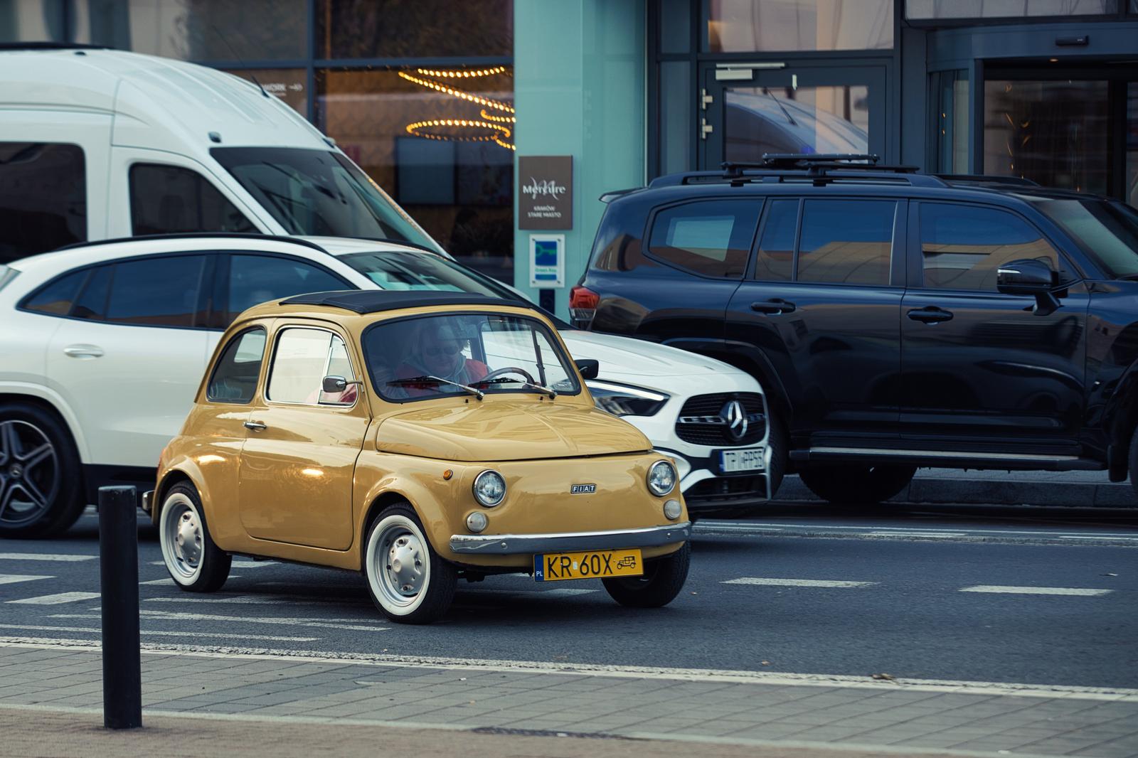 黄色いフィアット500がビルの前に駐車され、奥に白と黒の車が見える都市風景