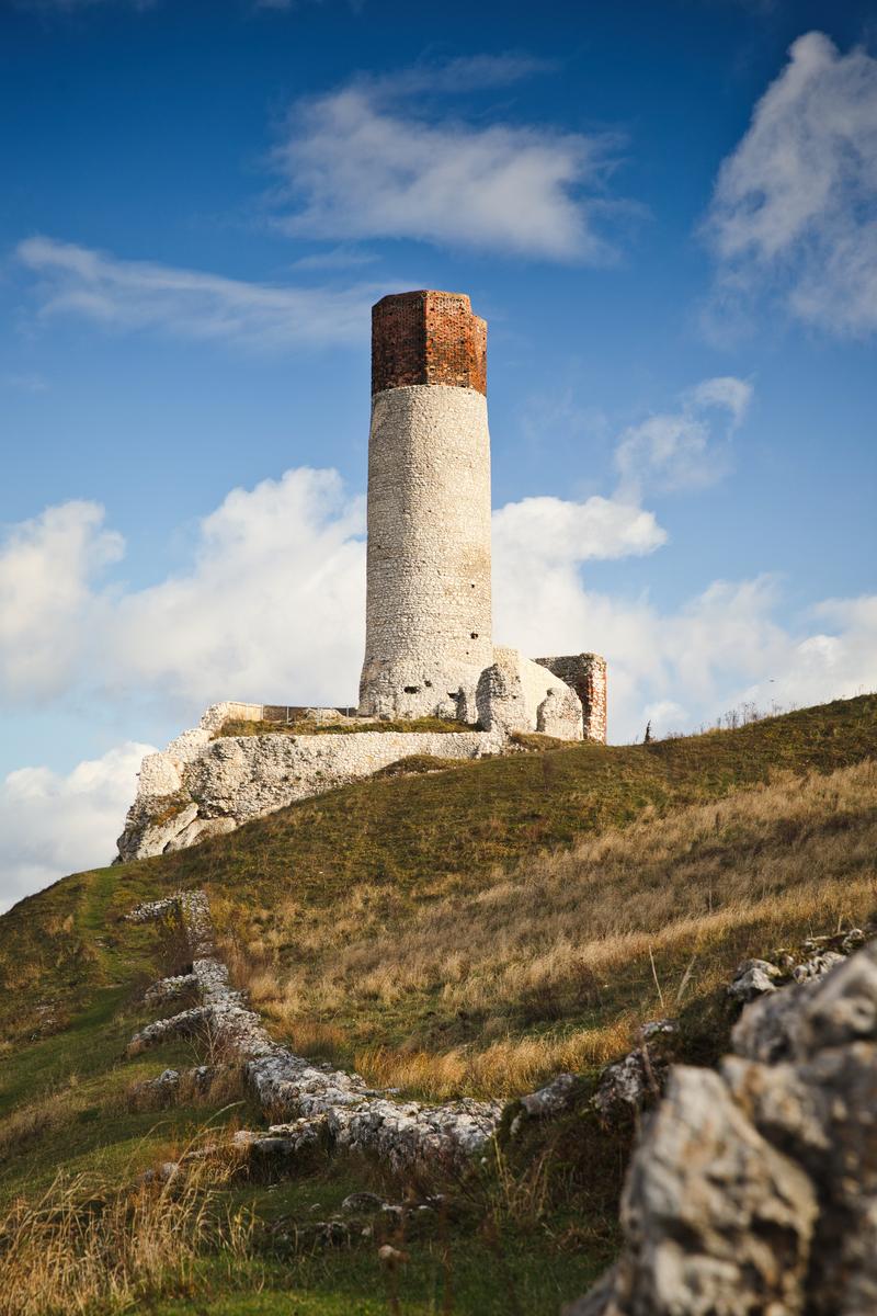 草の丘の上に立つ白い石造りの古い塔。上部は赤褐色のレンガ造り。周囲に遺構が残る