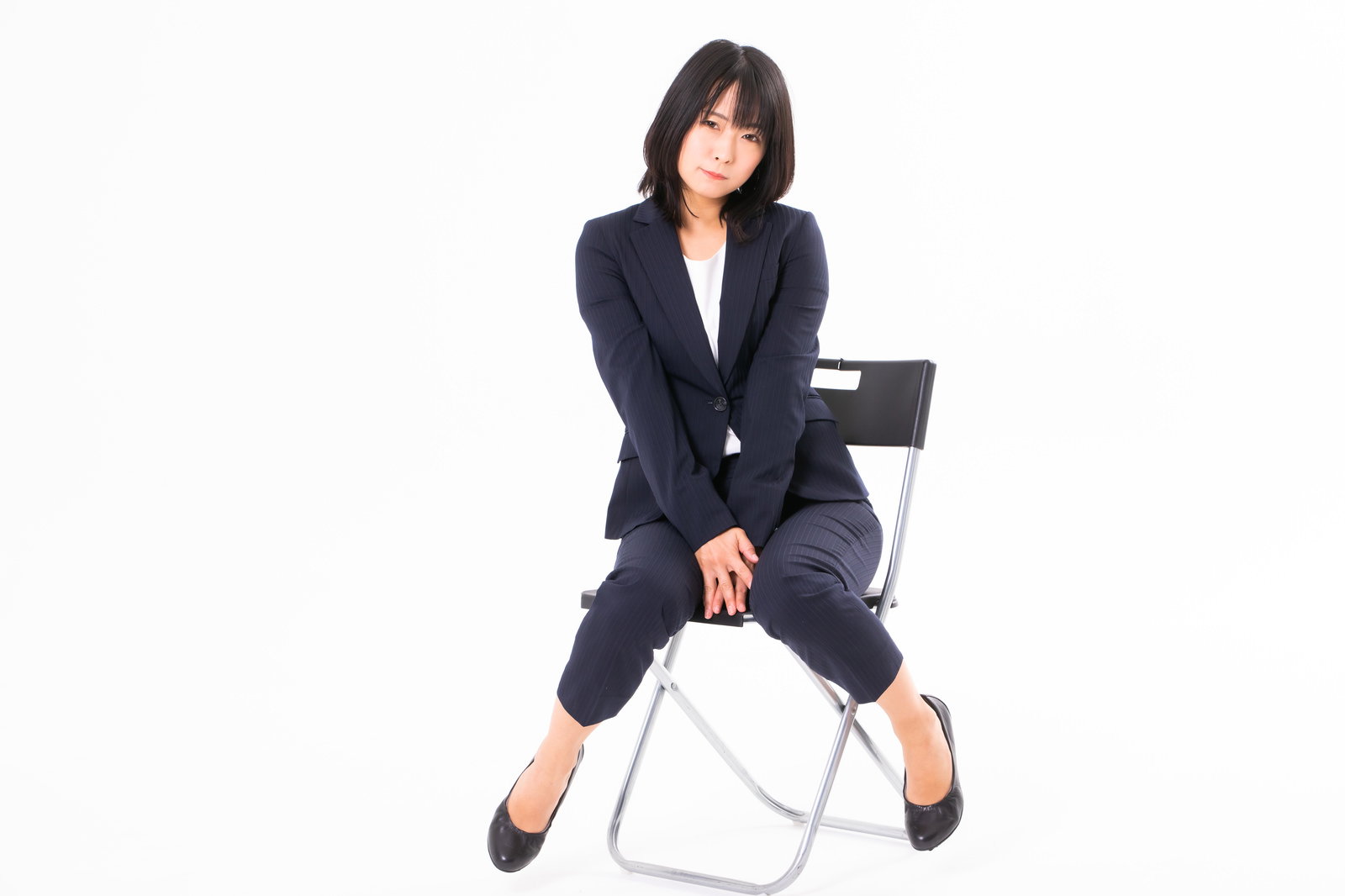 白い椅子に座りネイビーのスーツを着て首をかしげるポーズをとる女性社員［モデル：にゃるる］