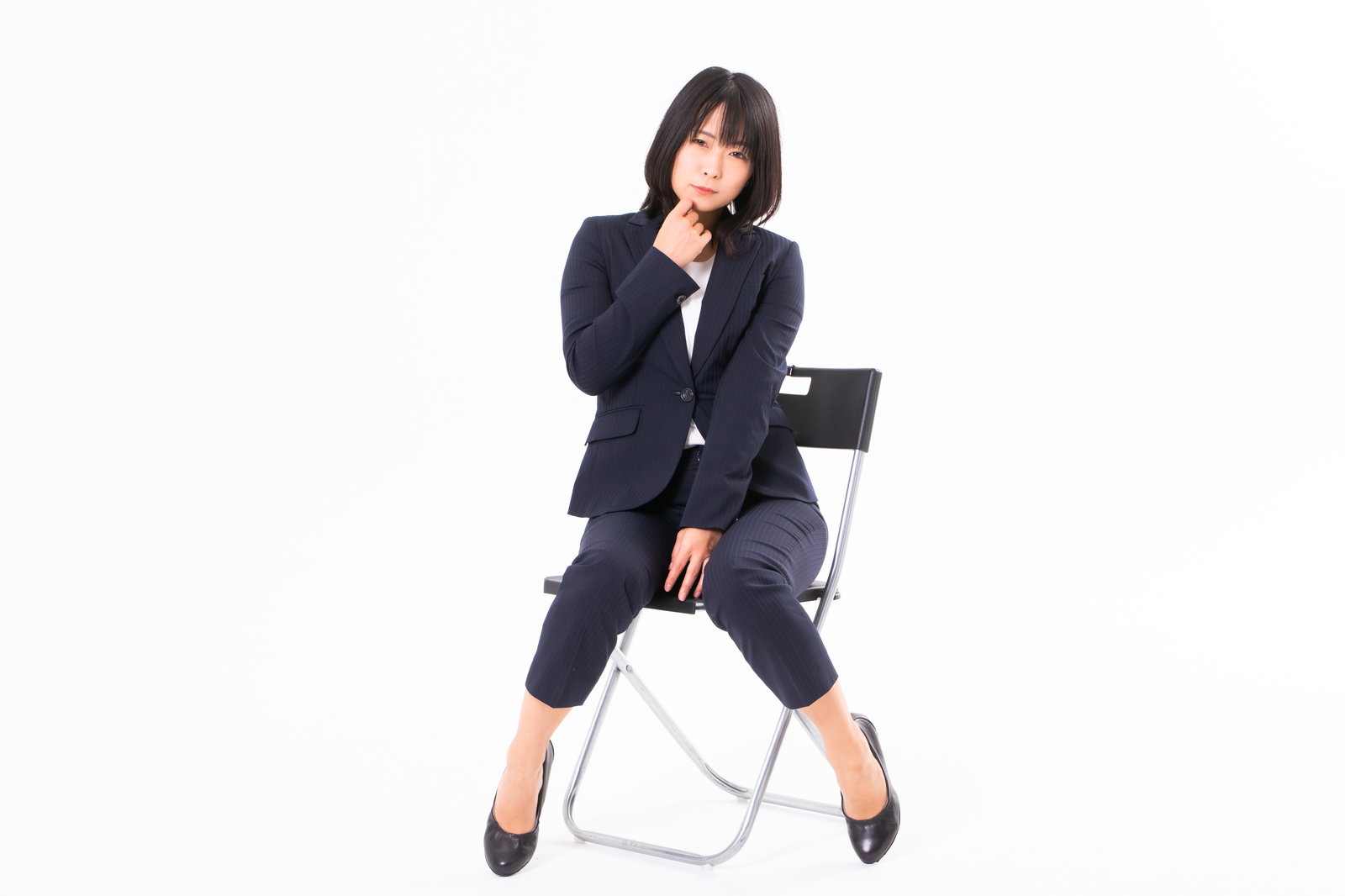 椅子に腰掛けて片足を組み、こちらを見つめる黒いスーツ姿の女性社員［モデル：にゃるる］