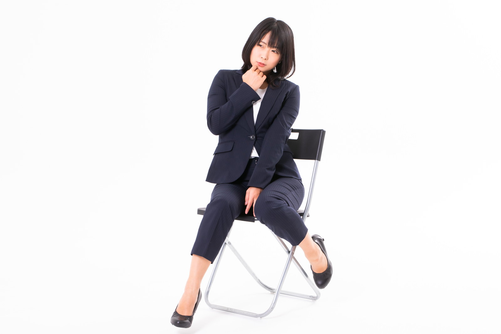 白い椅子に腰掛けて困惑した表情でモジモジする黒いスーツ姿の女性社員［モデル：にゃるる］