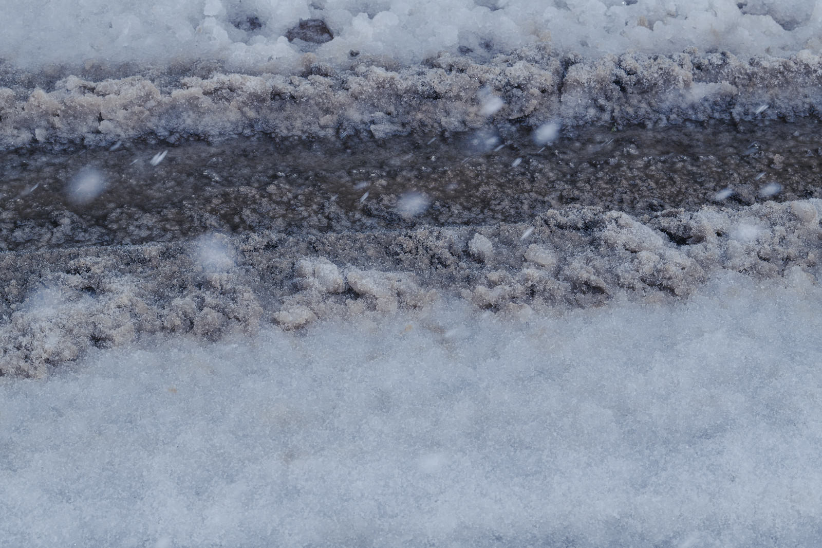 Winter road surface with multiple black tire tracks remaining on layered snow accumulation in white and dark gray tones