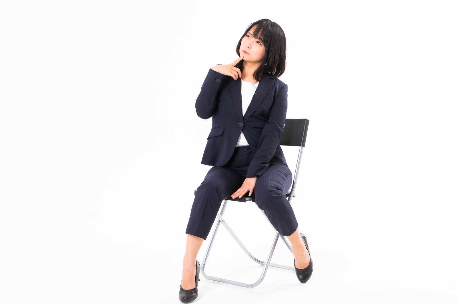 椅子に座って手を顎に当てて不思議そうな表情をする紺色スーツの女性社員［モデル：にゃるる］