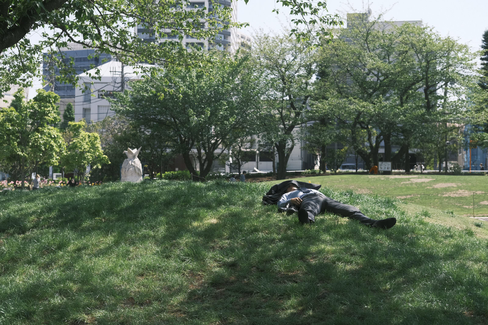 昼下がりの公園で緑の芝生に横になって昼寝をするスーツ姿のサラリーマン男性