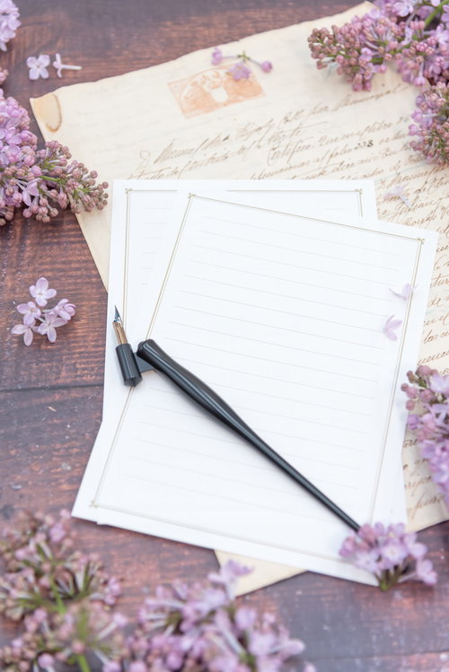 木のテーブルに散らばるライラックと記入前の便箋、万年筆