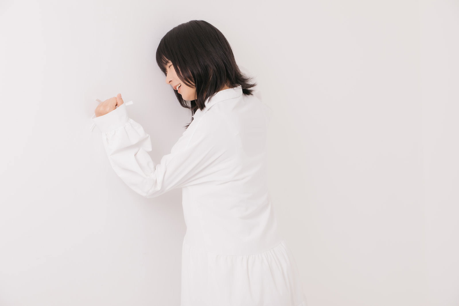 白い壁に向かって拳を当てて悔しがる白い服を着た女性の後ろ姿［モデル：にゃるる］