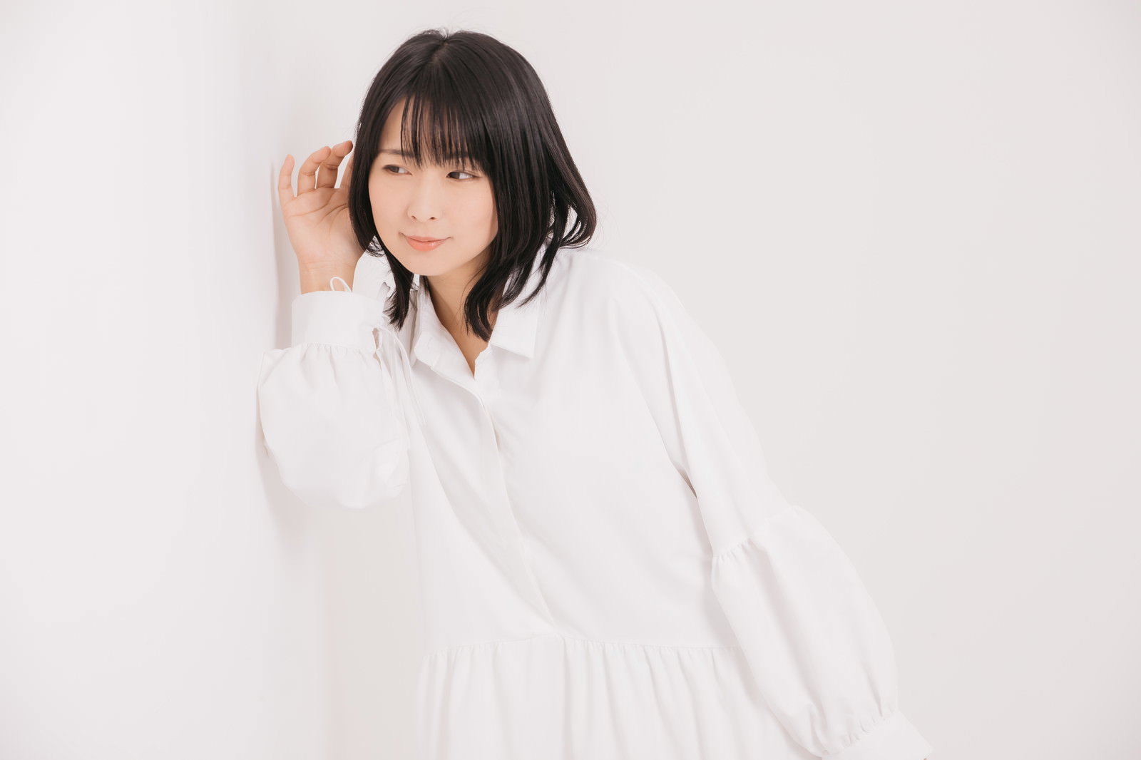 白いシャツの女性が壁に手と耳を当てて隣の音を聞いている様子［モデル：にゃるる］