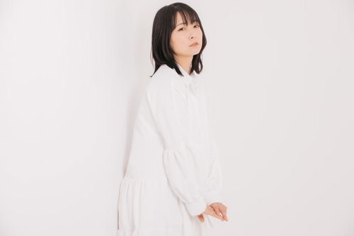 白い衣装で壁に背を向けてポージングする女性