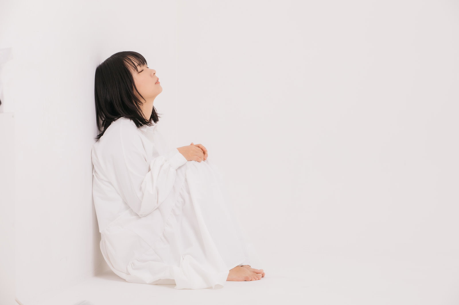 白い壁に寄りかかって座り込み、穏やかに寝落ちしている白い服の女性［モデル：にゃるる］