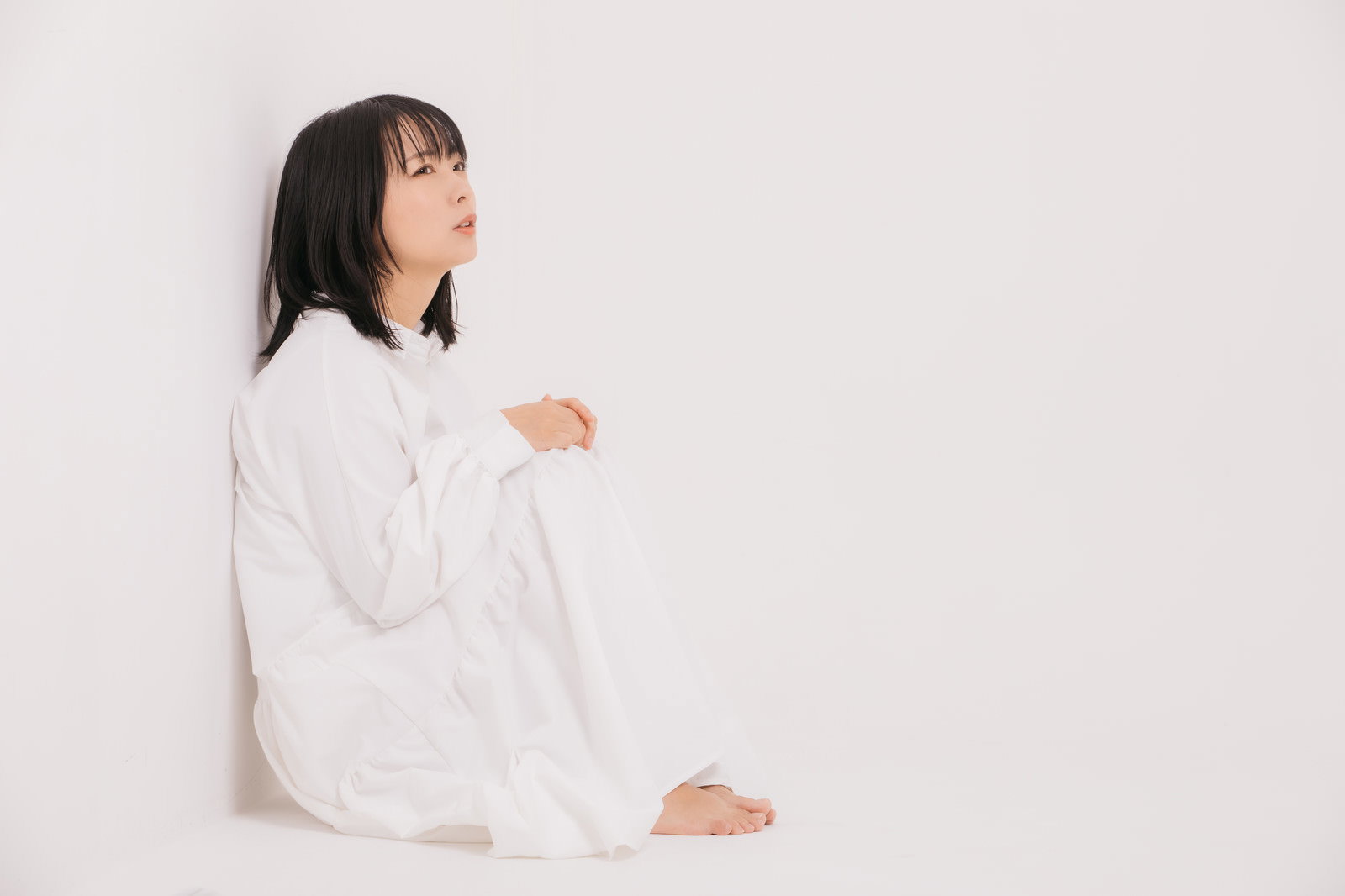 白い服を着た女性が壁際の隅っこに座り込み、膝を抱えてうつむいている様子［モデル：にゃるる］