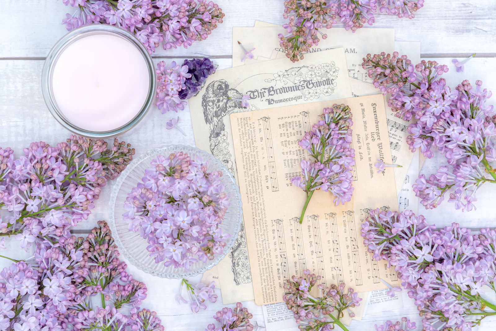 白いテーブルに散りばめられた紫のライラックの花と楽譜、キャンドルなどの雑貨