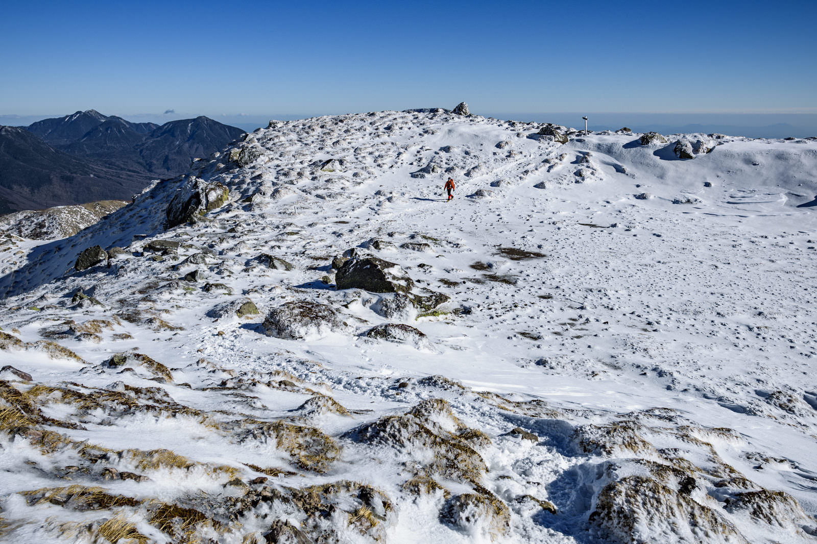 雪と岩が混在する日光白根山の山頂付近を歩く赤いウェアの登山者と青空