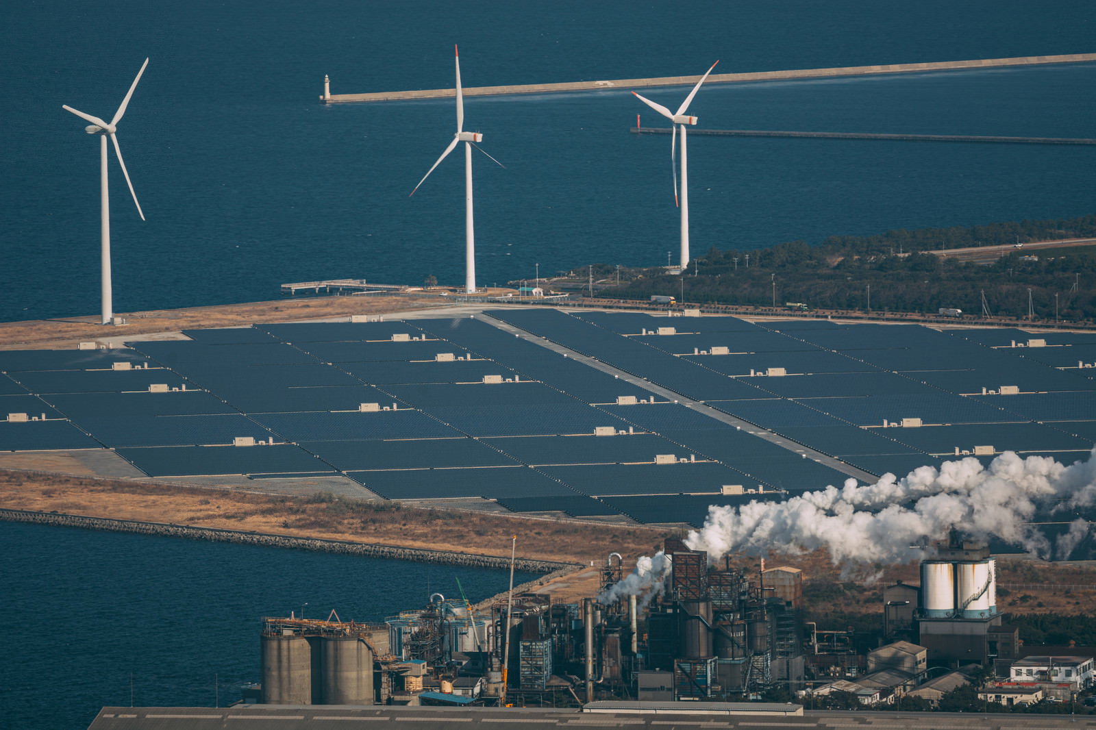 海岸沿いに設置された白い風力発電機と太陽光パネル、奥に工場が見える景観