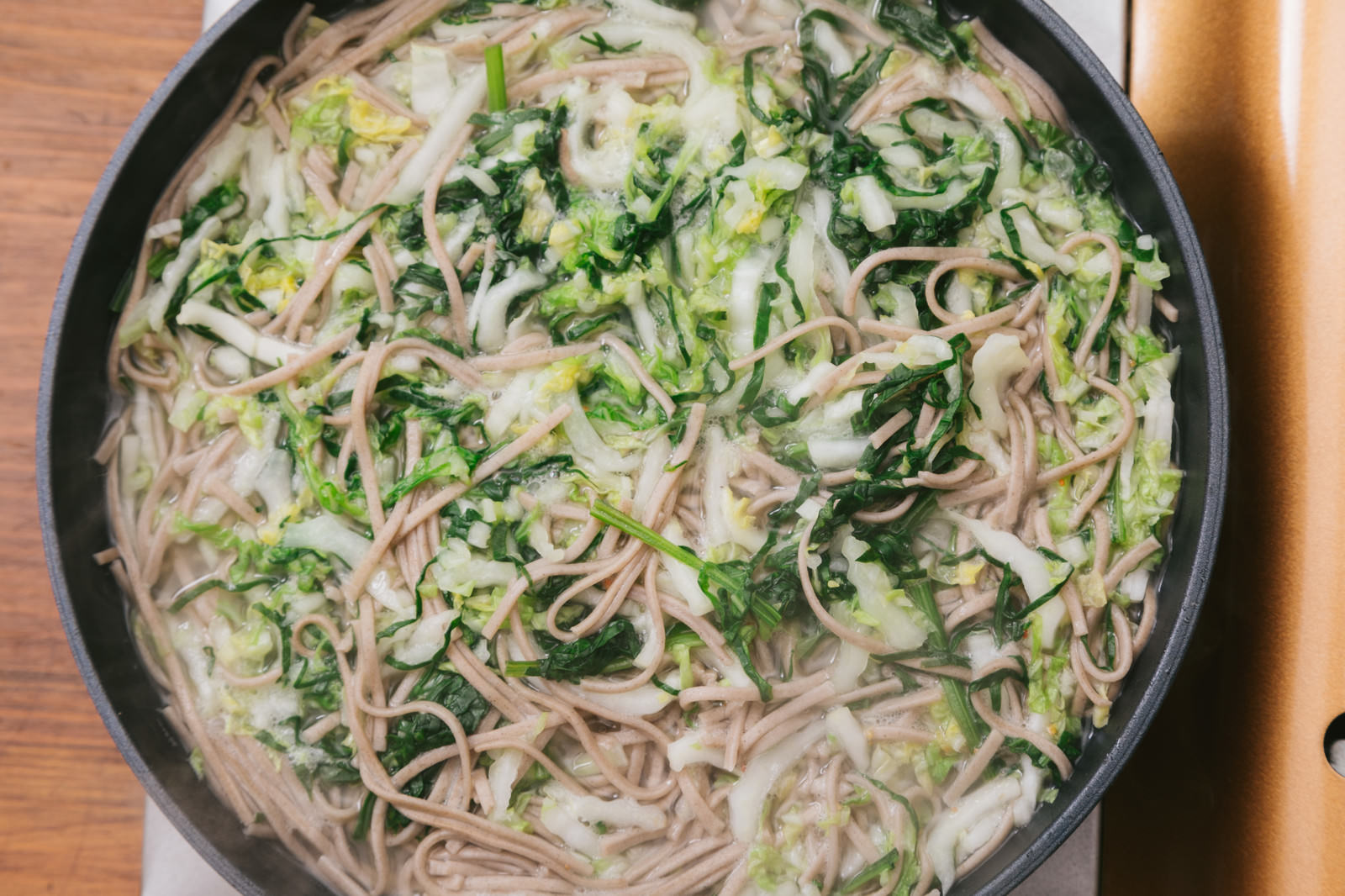黒い鍋に沸騰したお湯、そば麺、薄切り大根、白菜、ほうれん草が入った調理中の風景