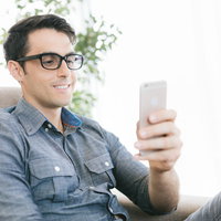 ソファーでスマホを見てニヤニヤする眼鏡の男性の写真