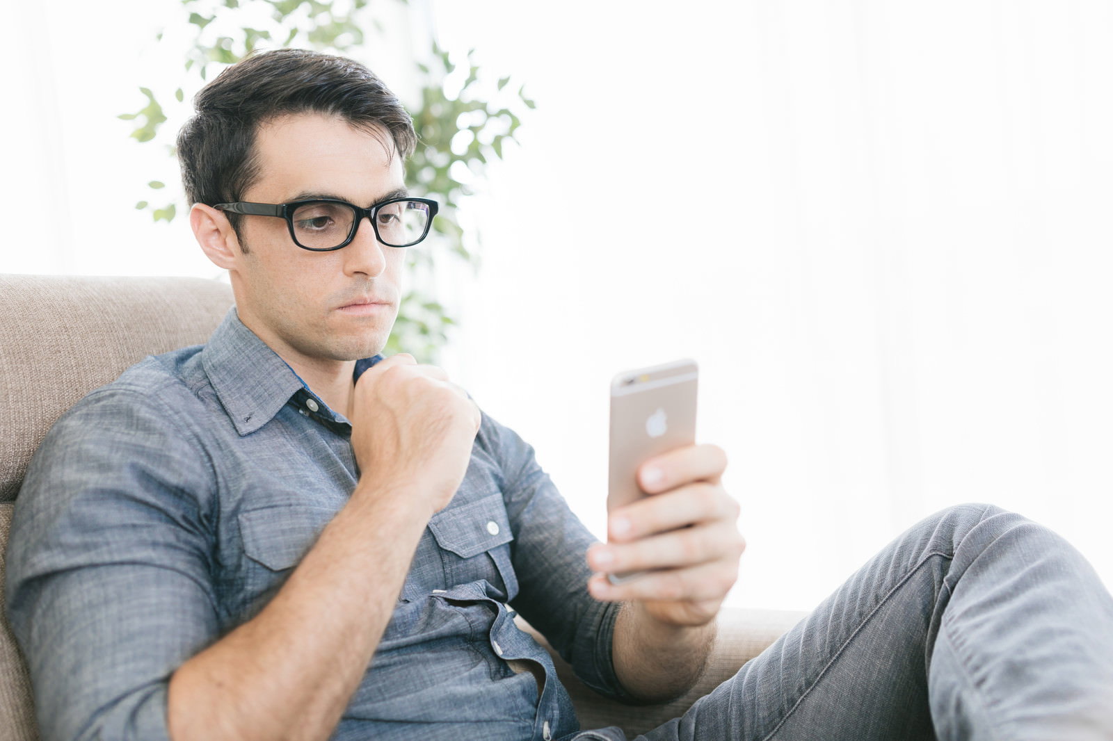 ソファに座りスマートフォンを見ながら顎に手を当てて考える眼鏡をかけた男性［モデル：アドリアーノ］
