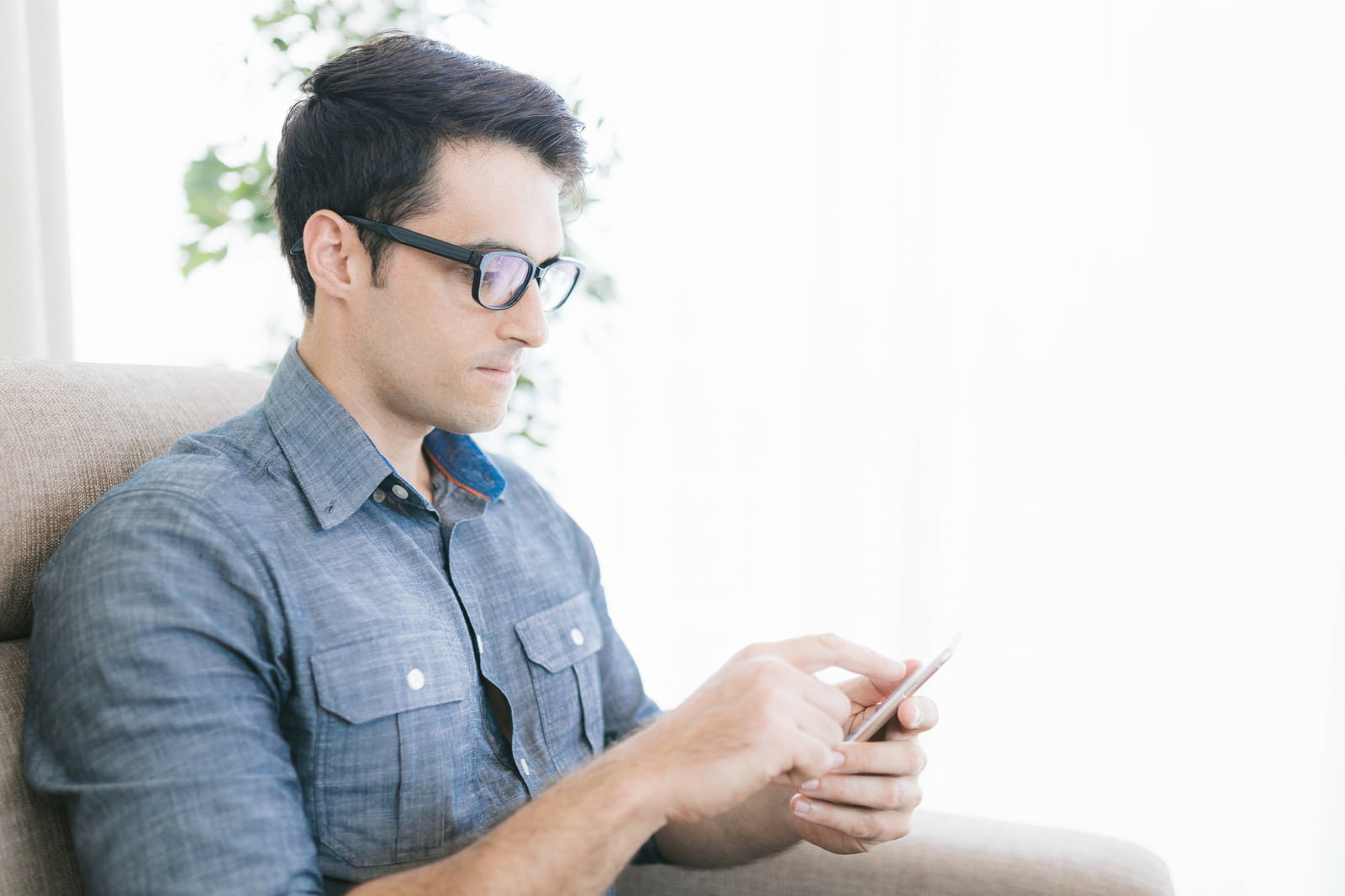 ソファに座ってスマートフォンを操作している眼鏡をかけた男性［モデル：アドリアーノ］