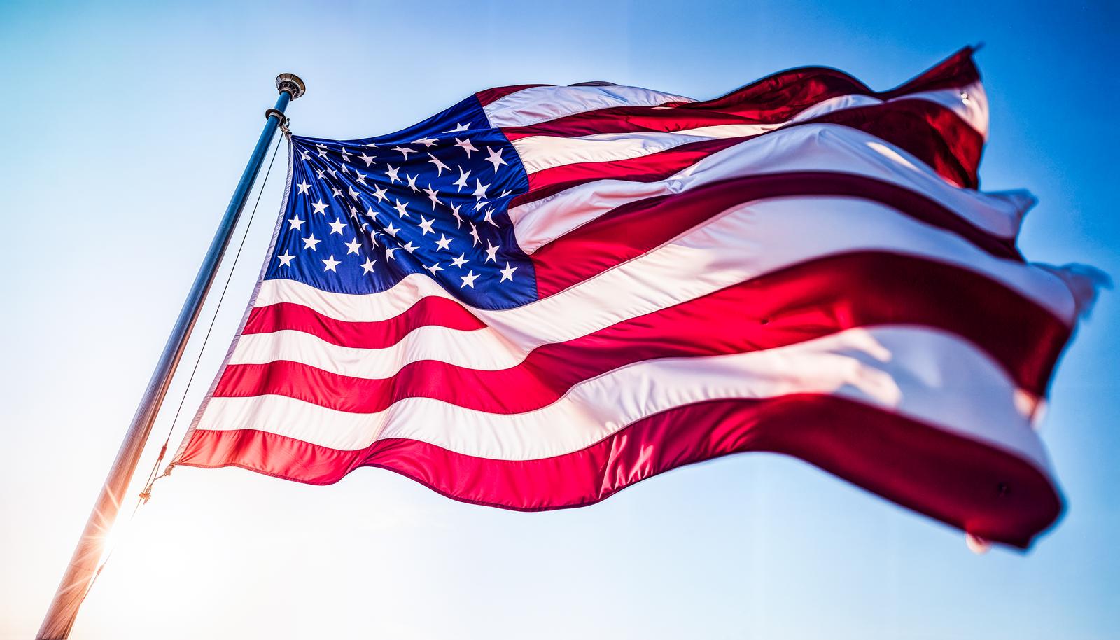 The Stars and Stripes flag raised on a pole against a blue sky background, waving significantly in the wind