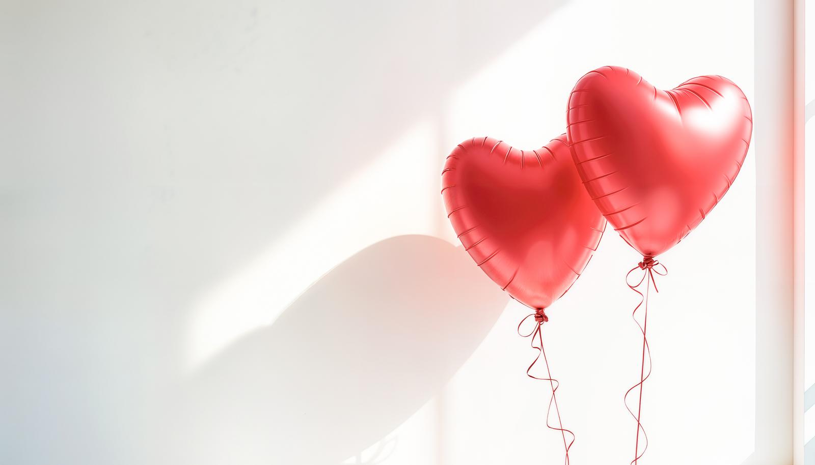 Two red heart-shaped foil balloons with curled ribbon ties floating against a plain white wall, soft natural light casting heart shadows.